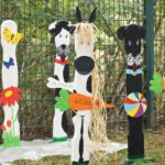 Geschmückte Holztiere mit Willkommensschildern stehen im Gras an einem Zaun.