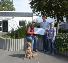 Drei Menschen und ein Hund halten einen großen Scheck vor dem Tierheim Siegen.