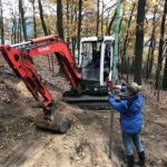 Zwei Personen arbeiten mit Werkzeugen und einem roten Bagger im Wald.