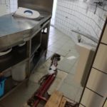 Zerbrochene Küchenfliesen legen rote Rohre in der Nähe der Metalltheke frei.