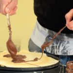 Verteilen von Schokolade auf einem Crêpe mit Löffeln, Glas in der Nähe.