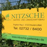 Gelbes Schild von Nitzsche Gartenbau mit Kontakten zwischen Grünpflanzen.