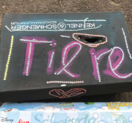 Schwarze Schachtel mit "Tiere!" in rosa Glitzer auf dem Boden.