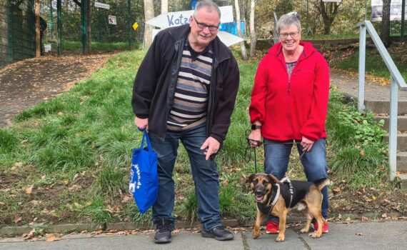 Zwei lächelnde Erwachsene mit einem glücklichen Hund an der Leine im Tierheim Siegen.
