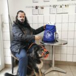 Frau mit Hund mit Maulkorb und blauer Tasche am Tisch in einem gekachelten Raum.