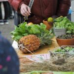 Person greift nach Ananas und Grünzeug am Markttisch im Freien.