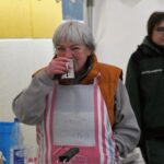 Ältere Frau in Schürze nippt an einem Becher; eine andere steht hinter ihr in der Küche.