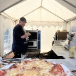 Mann im Zelt am Pizzaofen, frische Pizza auf dem Tablett davor.