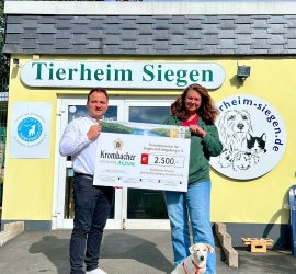 Zwei Personen mit einem großen Scheck im Tierheim Siegen; Hund zu ihren Füßen.