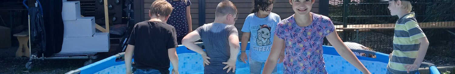 Sechs Kinder planschen an einem sonnigen Tag in einem blauen Hinterhofpool.