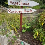 Drei deutsche Holzschilder weisen in verschiedene Richtungen durch einen Garten im Tierheim Siegen.