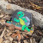 Gemalter Stein einer Cartoon-Schildkröte auf Gras, im Gartenmulch.