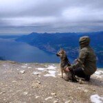 Person in grüner Jacke mit Hund auf einem Berggipfel, darunter ein See.