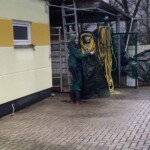 Person im Regenmantel hält Plane und gelbe Schläuche auf nassem Bürgersteig.