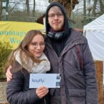 Draußen lächeln zwei Personen; eine hält eine kaufly-Karte neben einem gelben SWA-Banner.